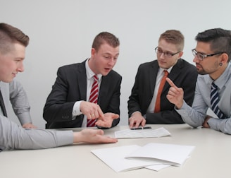 Four men in business attire are engaged in a discussion around a table. One man appears to be explaining something while pointing at some documents in front of them. The others are listening attentively, and one holds a pen and a mobile device. The setting is a plain office environment.