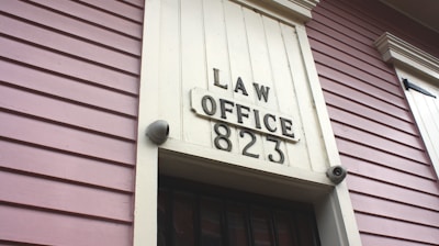 A close-up view of an exterior building wall with horizontal paneling painted in a soft pink color. The central focus is a large rectangular plaque stating 'LAW OFFICE 823' in bold letters. The paneling and design suggest a historical or vintage style.