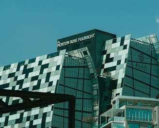 A modern, uniquely designed building with multiple reflective glass panels that create a checkered pattern. The upper parts display the names 'Norton Rose Fulbright' and another in Chinese characters. The structure exhibits a futuristic architectural style with sharp angles and a mix of straight and curved lines. In the foreground, additional buildings are visible with balconies and greenery, suggesting an urban setting.