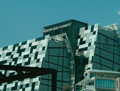 A modern, uniquely designed building with multiple reflective glass panels that create a checkered pattern. The upper parts display the names 'Norton Rose Fulbright' and another in Chinese characters. The structure exhibits a futuristic architectural style with sharp angles and a mix of straight and curved lines. In the foreground, additional buildings are visible with balconies and greenery, suggesting an urban setting.