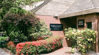 A brick building surrounded by lush greenery and vibrant flowers. The entrance is adorned with a sign reading 'Munro & Crawford, Barristers & Solicitors' and 'Notaries Public'. The address '5670 Yew Street' is displayed on a separate sign. There is a variety of foliage, including a large tree and shrubs with red and yellow flowers. A person is seated on a bench near the entrance, partially obscured by the plants.