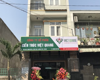 A multi-story building with a business on the ground floor displaying a sign for an architectural company. The facade includes balconies with railings. In front of the entrance, there is a table covered with a red cloth, adorned with decorative floral arrangements. Motorbikes are parked along the sidewalk and there are some potted plants on a ledge above the entrance.