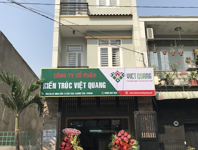 A multi-story building with a business on the ground floor displaying a sign for an architectural company. The facade includes balconies with railings. In front of the entrance, there is a table covered with a red cloth, adorned with decorative floral arrangements. Motorbikes are parked along the sidewalk and there are some potted plants on a ledge above the entrance.