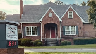 A traditional red brick building with a gabled roof and white-framed windows. The front entrance features an arched doorway and is flanked by well-maintained shrubs. A sign in the foreground reads 'Morris & Dean, Trial Lawyers & Counselors,' displaying the phone number and a digital clock showing 6:31.