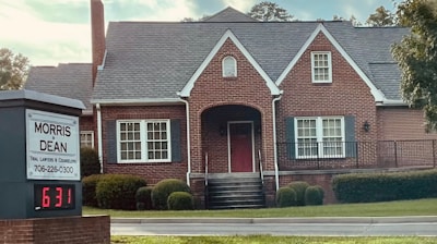 A traditional red brick building with a gabled roof and white-framed windows. The front entrance features an arched doorway and is flanked by well-maintained shrubs. A sign in the foreground reads 'Morris & Dean, Trial Lawyers & Counselors,' displaying the phone number and a digital clock showing 6:31.