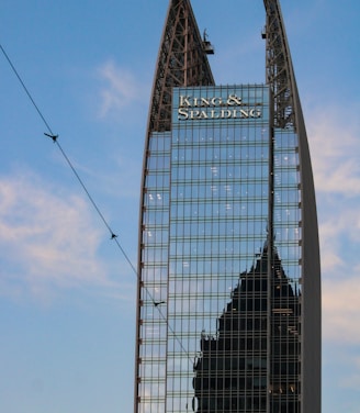 A modern skyscraper with a unique angular design features reflective glass panels. The name 'King & Spalding' is prominently displayed at the top, and another building’s silhouette is visible in the reflection. The blue sky and some thin clouds are visible in the background.