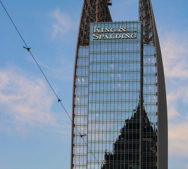 A modern skyscraper with a unique angular design features reflective glass panels. The name 'King & Spalding' is prominently displayed at the top, and another building’s silhouette is visible in the reflection. The blue sky and some thin clouds are visible in the background.